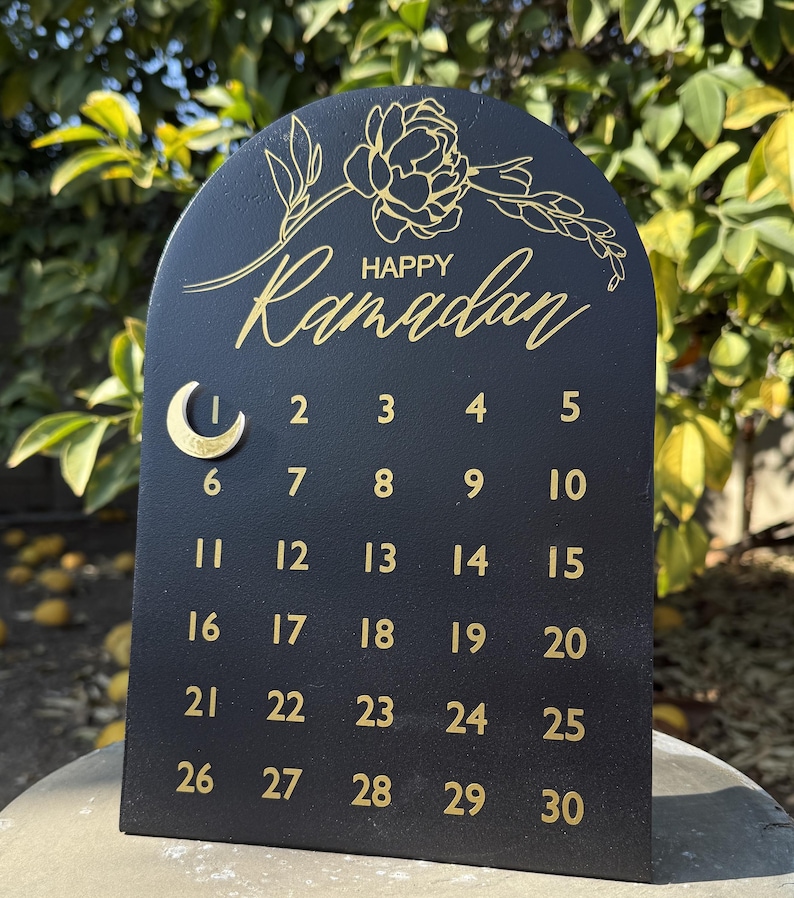 K&ouml;nnte beinhalten: Schwarzer Holzkalender mit goldener Beschriftung. Der Kalender ist bogenf&ouml;rmig und hat oben ein florales Design. Der Text "Happy Ramadan" steht oben auf dem Kalender. Die Zahlen 1 bis 30 sind in Reihen auf dem Kalender aufgelistet.