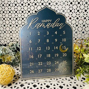 K&ouml;nnte beinhalten: Ein schwarzer Ramadan-Kalender aus Acryl mit goldenen Buchstaben und einem Halbmond. Der Kalender zeigt die Zahlen von 1 bis 30.