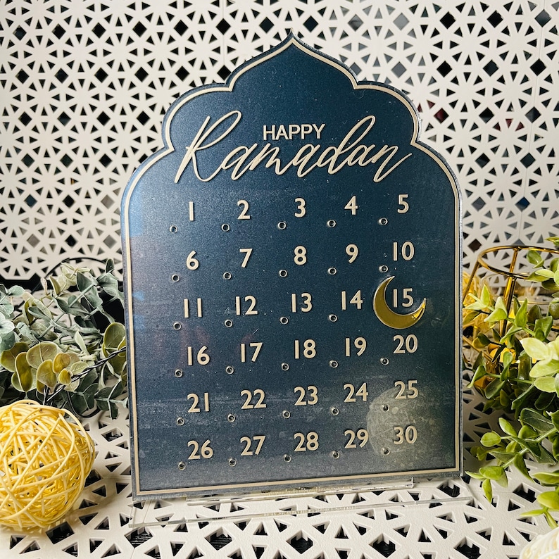 K&ouml;nnte beinhalten: Ein schwarzer Ramadan-Kalender aus Acryl mit goldenen Buchstaben und einem Halbmond. Der Kalender zeigt die Zahlen von 1 bis 30.