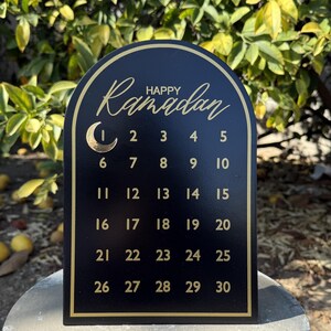 K&ouml;nnte beinhalten: Schwarzer Holzkalender mit goldenem Bogen und goldenen Buchstaben. Der Kalender zeigt "Frohes Ramadan" und die Zahlen von 1 bis 30.