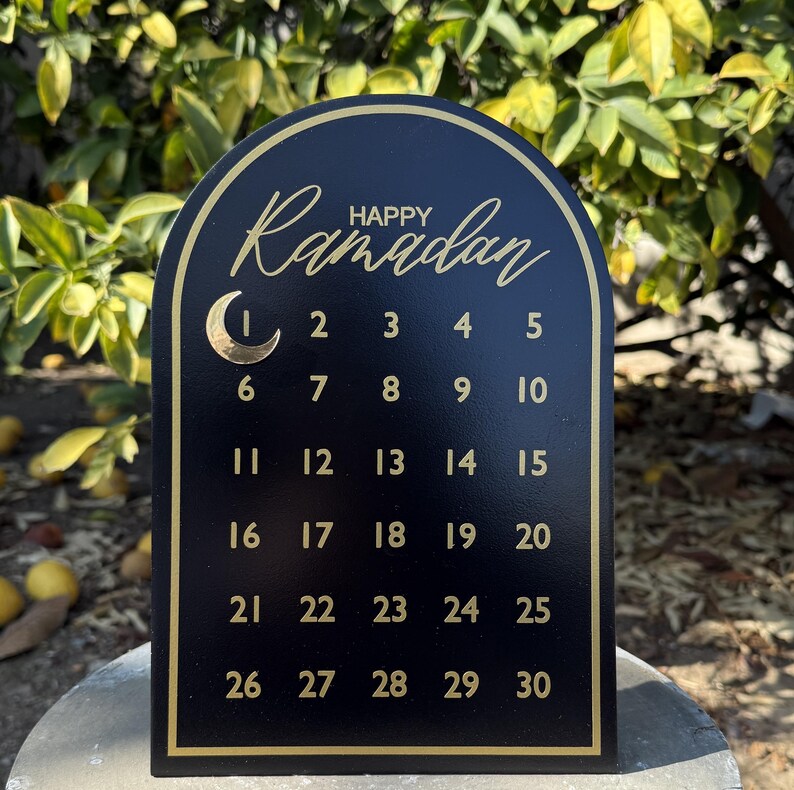 K&ouml;nnte beinhalten: Schwarzer Holzkalender mit goldenem Bogen und goldenen Buchstaben. Der Kalender zeigt "Frohes Ramadan" und die Zahlen von 1 bis 30.
