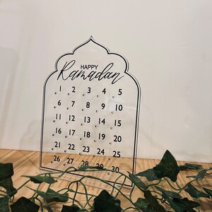 K&ouml;nnte beinhalten: Ein transparenter Ramadan-Kalender aus Acryl mit einem dekorativen Bogen oben. Der Kalender tr&auml;gt die Aufschrift "Happy Ramadan" oben und die Zahlen von 1 bis 30 in Schwarz.