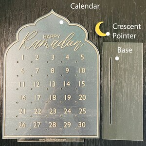 K&ouml;nnte beinhalten: Ein transparenter Ramadan-Kalender aus Acryl mit einem Halbmondzeiger und einem Sockel. Der Kalender zeigt die Zahlen von 1 bis 30 und den Text "Happy Ramadan".