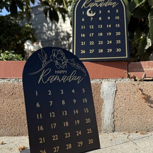 K&ouml;nnte beinhalten: Schwarzer Ramadan-Kalender aus Holz mit goldener Beschriftung und einem Halbmond-Design. Der Kalender zeigt ein florales Design und die Worte "Frohes Ramadan".