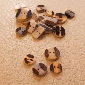 Puede incluir: Un conjunto de botones redondos de madera, cada uno con dos agujeros. Los botones tienen una base natural de color claro con detalles marrones y marrón oscuro. Están colocados sobre una superficie beige texturizada.