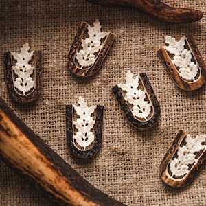 May include: Six intricately carved bone buttons with a floral design. The buttons are brown and white and have a U-shape. They are arranged on a hessian background.