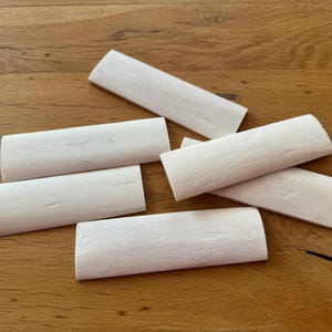 Könnte beinhalten: Sieben cremeweiße, rechteckige Knochenstücke mit abgerundeten Enden, angeordnet auf einer Holzoberfläche. Die Stücke haben eine leicht strukturierte Oberfläche. Die Holzmaserung ist im Hintergrund sichtbar. Die Stücke variieren in der Größe.