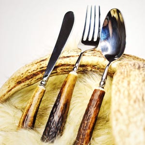 May include: A stainless steel knife, fork, and spoon set with handles made of brown and tan antler material. The silverware is displayed on a bed of faux fur and a piece of antler.