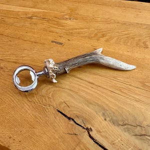 Peut inclure: Un décapsuleur doté d'un anneau en métal chromé poli et d'un manche en bois de cerf naturel brun et blanc. Le manche présente une surface texturée et organique avec une forme incurvée, fixée au mécanisme d'ouverture circulaire en métal.
