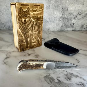 Peut inclure: Une boîte en bois avec une gravure détaillée de loup, un étui en cuir noir et un couteau pliant avec une lame à motifs et un manche en corne de cerf sont présentés sur une surface en marbre. Le couteau est ouvert, révélant son tranchant.