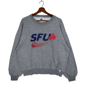 Puede incluir: Sudadera gris con un logotipo rojo y azul que dice "SFU" con una hoja de arce roja. El logotipo está en un swoosh blanco con una franja roja.