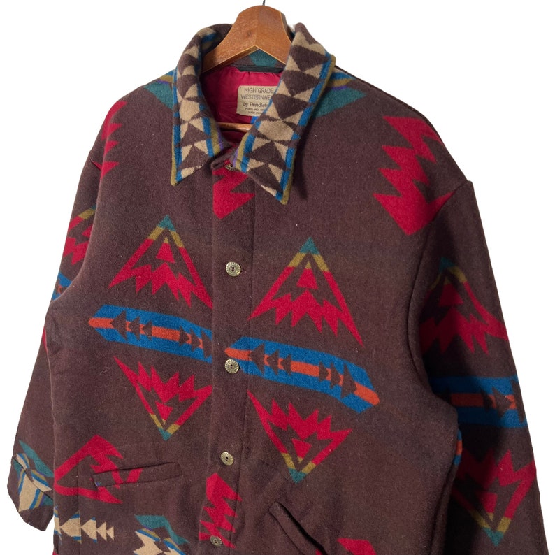 May include: A brown wool jacket with a geometric pattern in red, blue, and tan. The jacket has a collared neckline and button closure. The collar features a unique geometric design. The jacket is labeled "HIGH GRADE WESTERNWEAR by Pendleton".