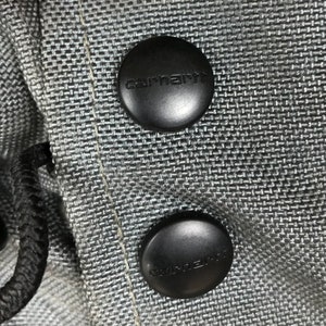 May include: Close-up of two black buttons with the word "carhartt" embossed on them. The buttons are attached to a gray fabric.