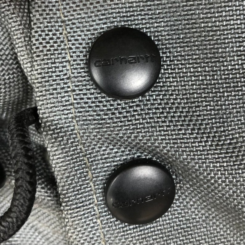 May include: Close-up of two black buttons with the word "carhartt" embossed on them. The buttons are attached to a gray fabric.