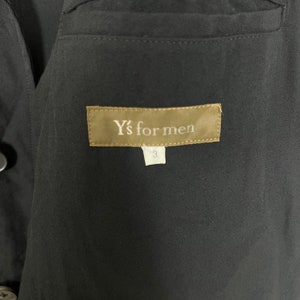 Op de afbeelding: Close-up van een zwart kledingstuk met een label met de tekst "Y's for men" in witte letters op een bruine achtergrond. Onder het label staat een wit label met het nummer "3". Het kledingstuk heeft een getextureerde uitstraling.