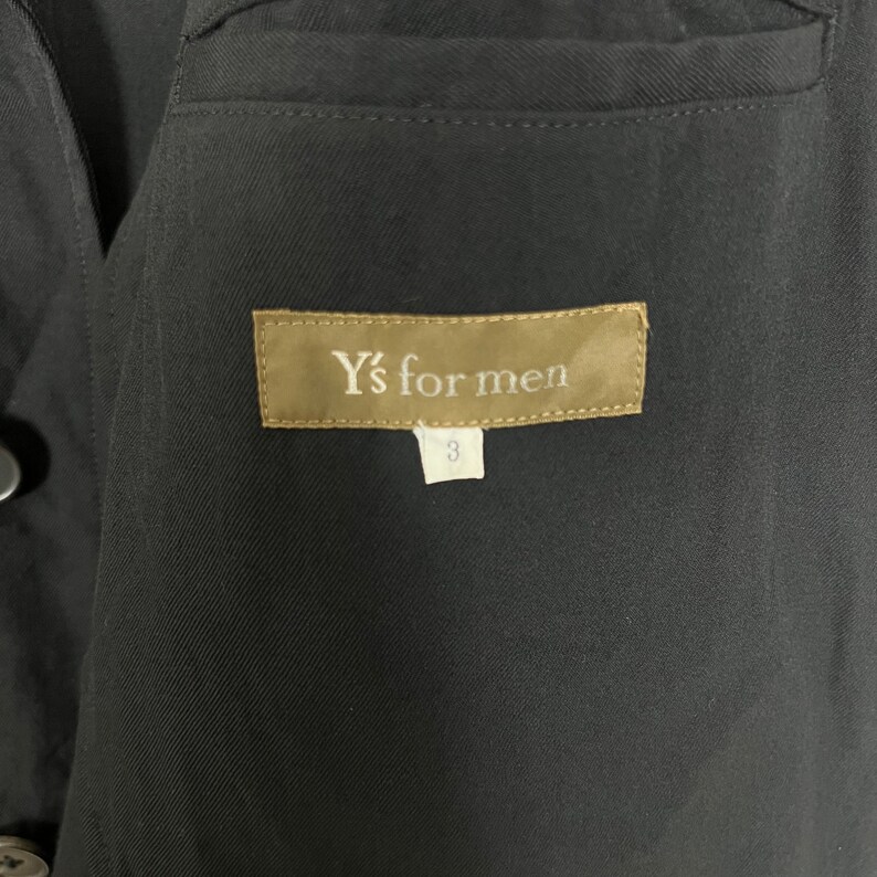 Op de afbeelding: Close-up van een zwart kledingstuk met een label met de tekst "Y's for men" in witte letters op een bruine achtergrond. Onder het label staat een wit label met het nummer "3". Het kledingstuk heeft een getextureerde uitstraling.