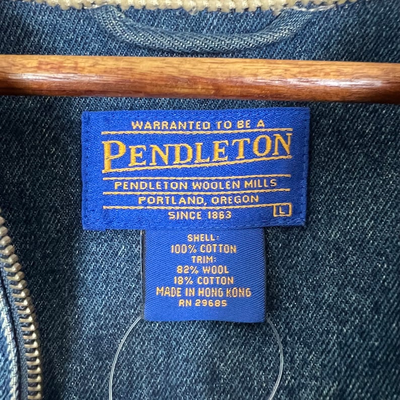 Pode incluir: Close-up de uma jaqueta jeans azul com uma etiqueta Pendleton. A etiqueta &eacute; azul com texto dourado e inclui o nome da marca, localiza&ccedil;&atilde;o e composi&ccedil;&atilde;o do tecido. A jaqueta tem um z&iacute;per e um forro claro.