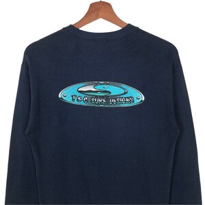 Pu&ograve; includere: Una maglietta a maniche lunghe blu navy con una grafica di un simbolo yin-yang all'interno di un cerchio con il testo "TSC Surf Designs".