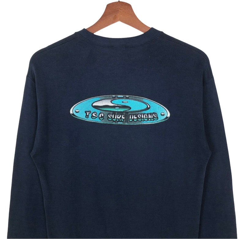Pu&ograve; includere: Una maglietta a maniche lunghe blu navy con una grafica di un simbolo yin-yang all'interno di un cerchio con il testo "TSC Surf Designs".
