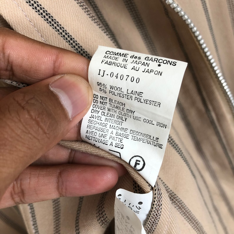 K&ouml;nnte beinhalten: Ein beige-braunes, gestreiftes Stofflabel mit dem Text "COMME des GARCONS" und "MADE IN JAPAN" in Schwarz. Das Etikett enth&auml;lt auch den Text "IJ-040700", "95% WOOL LAINE", "5% POLYESTER POLYESTER" und Pflegehinweise in Schwarz.