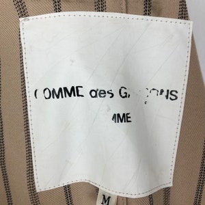 K&ouml;nnte beinhalten: Ein wei&szlig;es Etikett mit schwarzem Text, der "COMME des GARCONS" und "ME" lautet, auf einem braunen und schwarz gestreiften Stoffhintergrund. Das Etikett hat auch ein wei&szlig;es Etikett mit einem schwarzen "M" darauf.