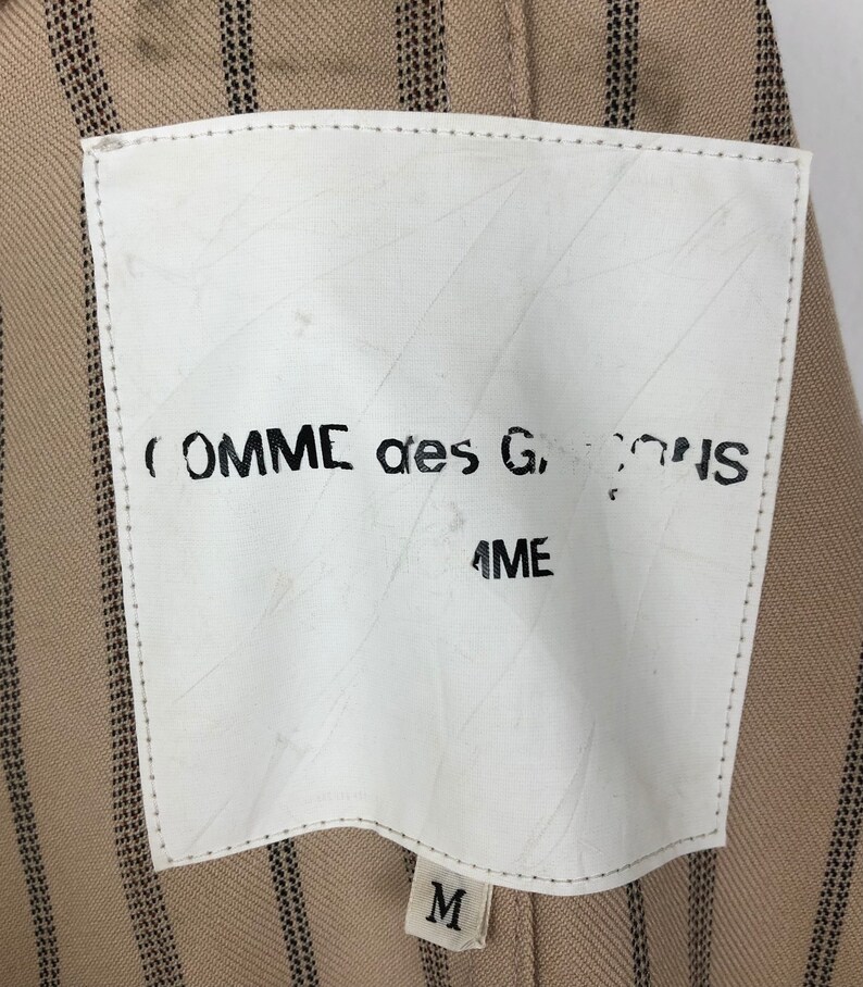 K&ouml;nnte beinhalten: Ein wei&szlig;es Etikett mit schwarzem Text, der "COMME des GARCONS" und "ME" lautet, auf einem braunen und schwarz gestreiften Stoffhintergrund. Das Etikett hat auch ein wei&szlig;es Etikett mit einem schwarzen "M" darauf.