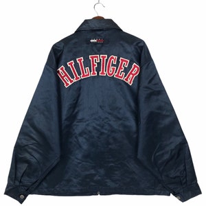 May include: A navy blue satin jacket with a red and white "HILFIGER" logo on the back. The jacket has a zipper closure and a button-up collar.
