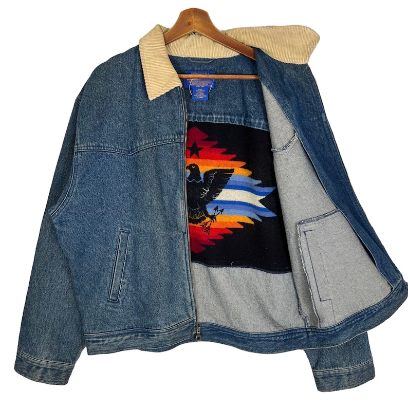 Pode incluir: Casaco de ganga azul com gola de veludo cotel&ecirc;. O casaco apresenta um forro estampado colorido com um desenho de &aacute;guia. Possui fecho de correr e dois bolsos frontais.