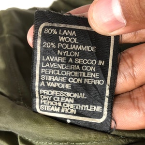 Pu&ograve; includere: Primo piano di un'etichetta di abbigliamento nera con testo bianco. L'etichetta indica la composizione del tessuto come 80% lana e 20% nylon, con istruzioni per il lavaggio a secco e la stiratura a vapore. L'etichetta &egrave; contro un tessuto verde.
