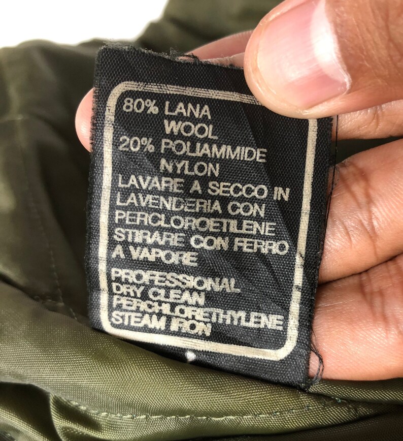 Pu&ograve; includere: Primo piano di un'etichetta di abbigliamento nera con testo bianco. L'etichetta indica la composizione del tessuto come 80% lana e 20% nylon, con istruzioni per il lavaggio a secco e la stiratura a vapore. L'etichetta &egrave; contro un tessuto verde.