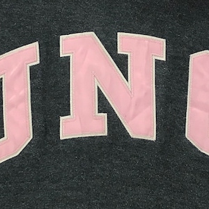 May include: Pink "UNC" lettering in a collegiate-style font on a dark gray background.