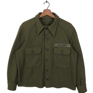 Puede incluir: Una camisa de lana verde con cierre de botones y dos bolsillos de solapa. La camisa tiene un parche en el bolsillo derecho que dice "U.S. ARMY".