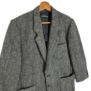 Puede incluir: Una blazer de tweed de espiga gris y negro con solapa de muesca, cierre de tres botones y dos bolsillos delanteros. La blazer est&aacute; colgada de una percha de madera. La etiqueta dentro de la blazer dice "tricot COMME des GAR&Ccedil;ONS."