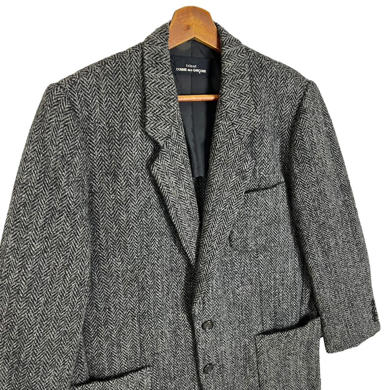 Puede incluir: Una blazer de tweed de espiga gris y negro con solapa de muesca, cierre de tres botones y dos bolsillos delanteros. La blazer est&aacute; colgada de una percha de madera. La etiqueta dentro de la blazer dice "tricot COMME des GAR&Ccedil;ONS."