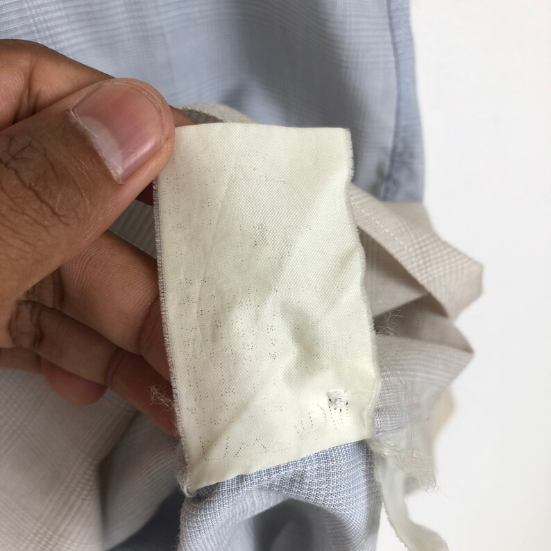 Op de afbeelding: Een close-up van een wit stoffen label met een klein gaatje in het midden. Het label is bevestigd aan een lichtblauwe en wit gestreepte stof.