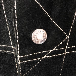 Puede incluir: Primer plano de una tela de ante negro con costuras blancas. Un bot&oacute;n de metal plateado con la palabra "Diesel" y una estrella es visible en el centro.