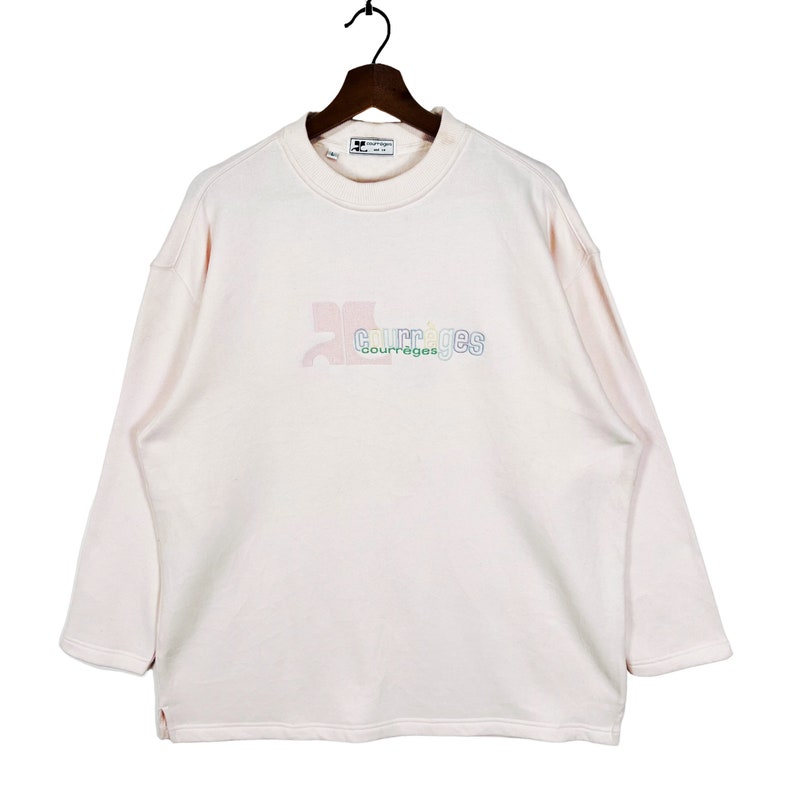 May include: A pale pink Courr&egrave;ges sweatshirt with long sleeves, hanging on a wooden hanger. The brand's logo is embroidered in a gradient of colours across the chest. The sweatshirt has a crew neck and a relaxed fit.