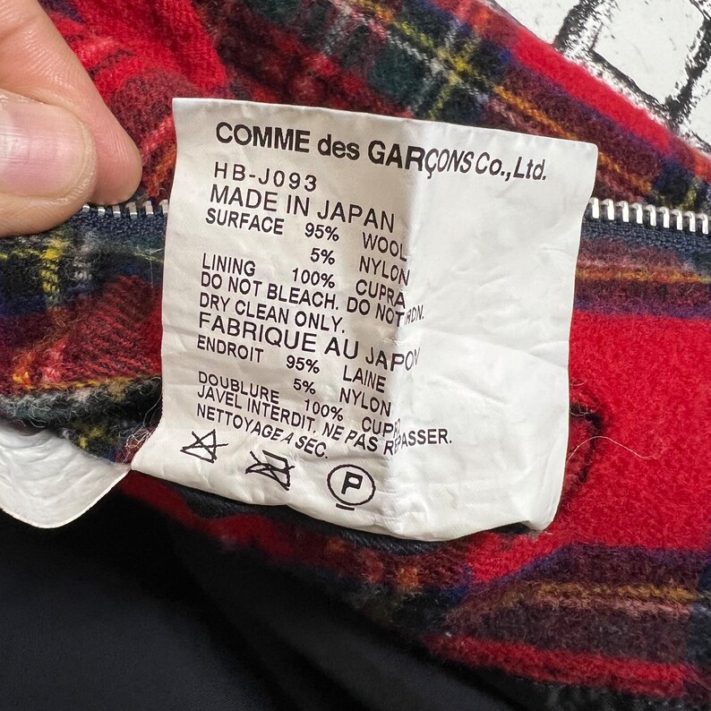 May include: White tag with black text detailing the composition of a garment. The tag reads "COMME des GAR&Ccedil;ONS Co., Ltd. HB-J093 MADE IN JAPAN SURFACE 95% WOOL 5% NYLON LINING 100% CUPRA DO NOT BLEACH. DO NOT IRON. DRY CLEAN ONLY. FABRIQUE AU JAPON ENDROIT 95% LAINE DOUBLURE 100% CUPRA 5% NYLON JAVEL INTERDIT. NE PAS REPASSER. NETTOYAGE A SEC. P"