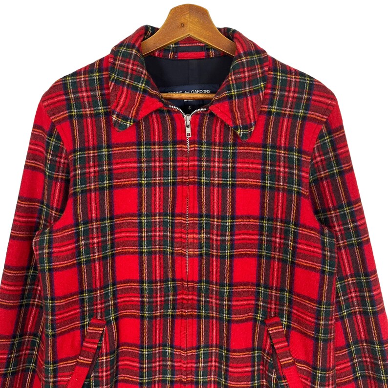 May include: Red and black plaid jacket with a zipper closure. The jacket has a collar and two pockets. The label on the jacket reads "Comme des Gar&ccedil;ons."
