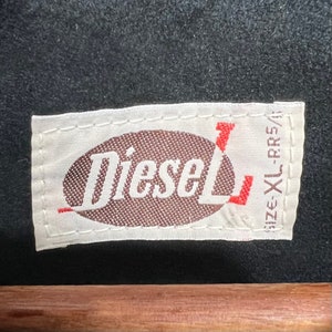 Puede incluir: Etiqueta blanca con un &oacute;valo marr&oacute;n que contiene la palabra "Diesel" en texto blanco. La etiqueta tiene un texto rojo "Size XL-CRS" en el lado derecho.