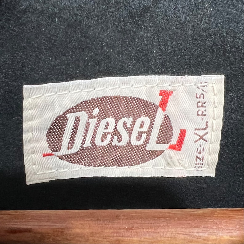 Puede incluir: Etiqueta blanca con un &oacute;valo marr&oacute;n que contiene la palabra "Diesel" en texto blanco. La etiqueta tiene un texto rojo "Size XL-CRS" en el lado derecho.