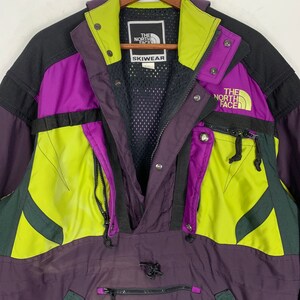 May include: A purple, black, green, and yellow ski jacket with a zipper closure and a black mesh lining. The jacket has a The North Face logo on the right side and a "Skiwear" label on the left side.