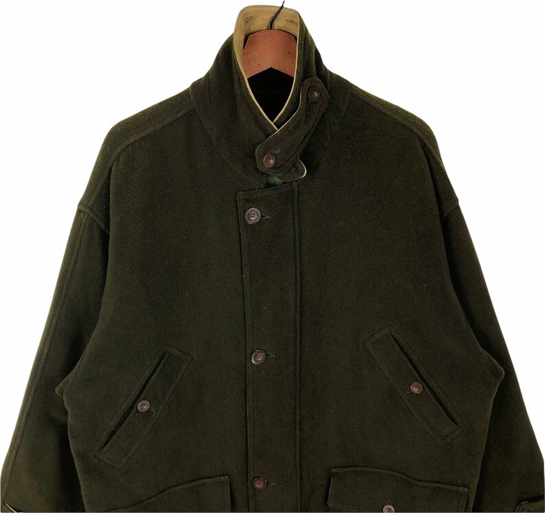 Pu&ograve; includere: Un cappotto di lana verde scuro con collo alto e chiusura con bottoni. Il cappotto ha due tasche anteriori e una fodera marrone chiaro visibile sul colletto. Uno stile classico, adatto al clima freddo.