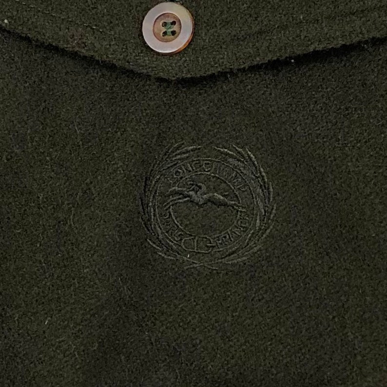 Pu&ograve; includere: Primo piano di un tessuto verde scuro con un bottone e una patta. La patta presenta un emblema ricamato con un cavallo e le parole "LONGCHAMP PARIS FRANCE" in un design circolare. Il bottone &egrave; marrone chiaro.