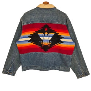 May include: A blue denim jacket with a colourful Native American inspired pattern on the back. The pattern features a red, orange, yellow, blue, and white striped design with a black eagle in the centre.