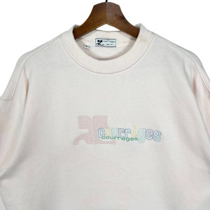 May include: Pale pink Courr&egrave;ges sweatshirt with a crew neck and long sleeves. The brand logo is embroidered in pastel colours on the front. The sweatshirt is hanging on a wooden hanger.