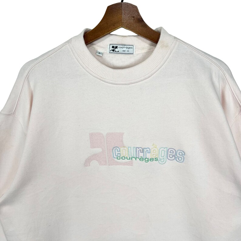 May include: Pale pink Courr&egrave;ges sweatshirt with a crew neck and long sleeves. The brand logo is embroidered in pastel colours on the front. The sweatshirt is hanging on a wooden hanger.