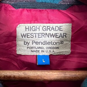 May include: Close-up of a label from a Pendleton Westernwear jacket. The label is tan with black text that reads "HIGH GRADE WESTERNWEAR by Pendleton PORTLAND, OREGON MADE IN U.S.A." A blue tag with a white "L" is attached. The jacket lining is burgundy.