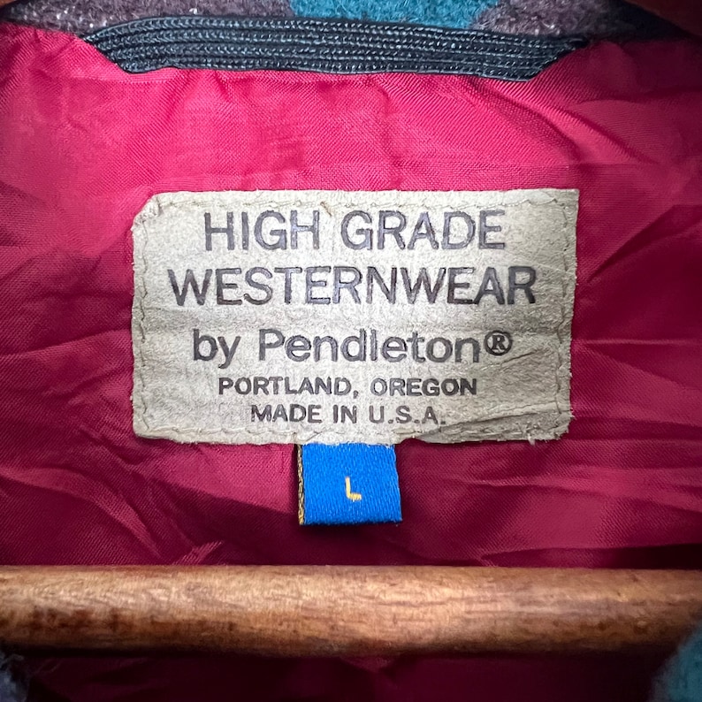 May include: Close-up of a label from a Pendleton Westernwear jacket. The label is tan with black text that reads "HIGH GRADE WESTERNWEAR by Pendleton PORTLAND, OREGON MADE IN U.S.A." A blue tag with a white "L" is attached. The jacket lining is burgundy.