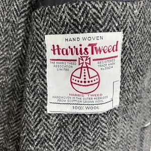 Puede incluir: Primer plano de una tela de tweed gris y negro con dise&ntilde;o de espiga. Una etiqueta blanca con texto rojo dice "Harris Tweed", con detalles sobre su origen y que est&aacute; tejida a mano con lana escocesa.